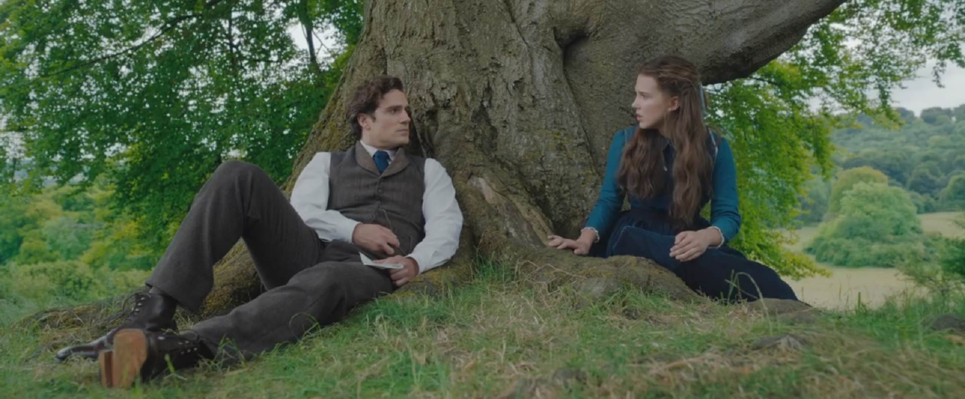 بابی میلی براون در نقش انولا و هنری کویل در نقش شرلوک هولمز با بک گراند درخت تنومند، شاخه‌ها، چمن و تپه‌ی سبز در فیلم Enola Holmes