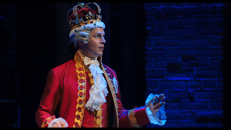 پادشاه انگلستان در نمایش موزیکال همیلتون با لباس قرمز و سفید سلطنتی و تاج بزرگ