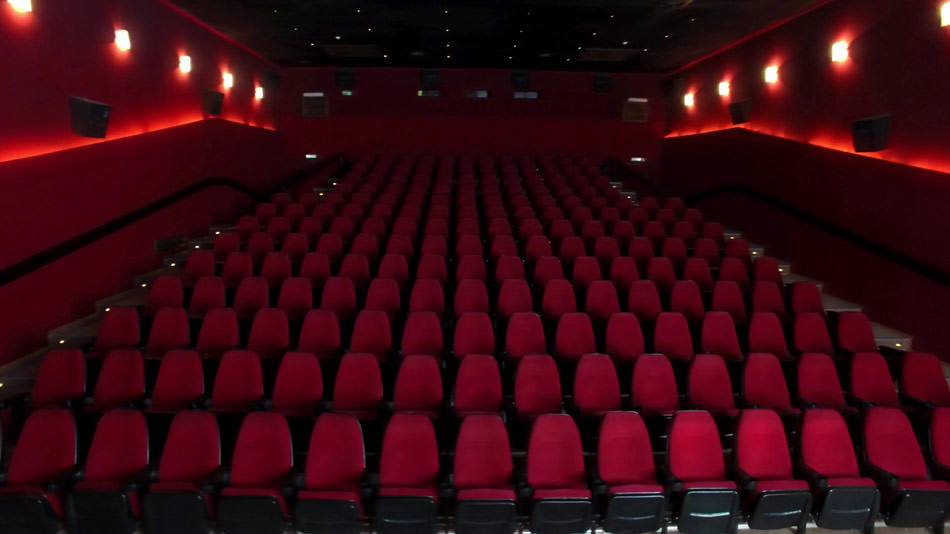 سالن خالی و بزرگ سینما با چند ردیف صندلی قرمز خالی و چراغ های روشن