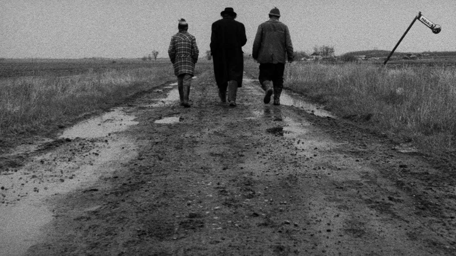 سه نفر در حال قدم زدن در جاده گلی فیلم سیاه و سفید Satantango