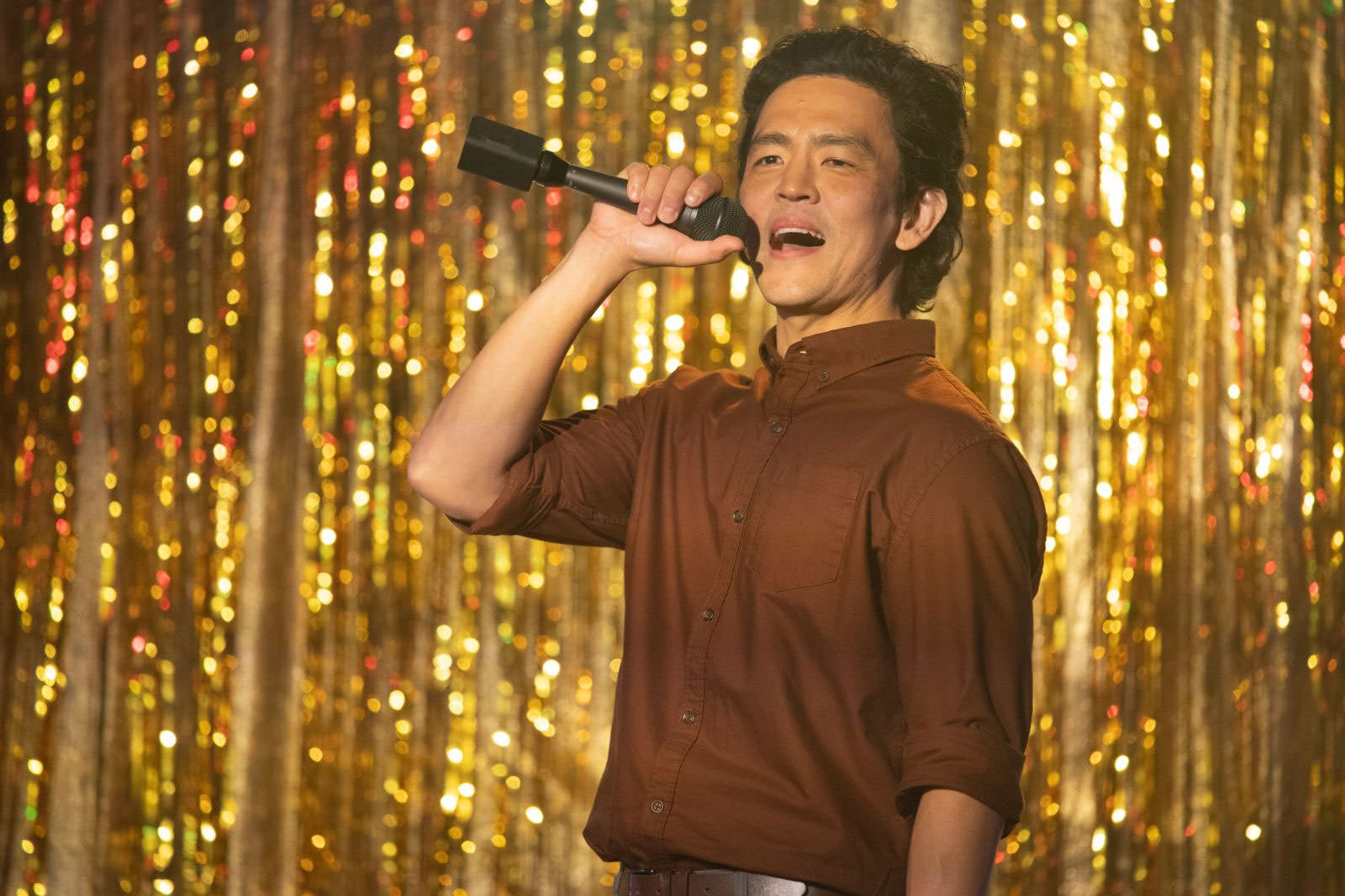 John Cho, Don't Make Me Go filminden bir sahnede arkasında altın bir fon varken elinde mikrofonla şarkı söylüyor.
