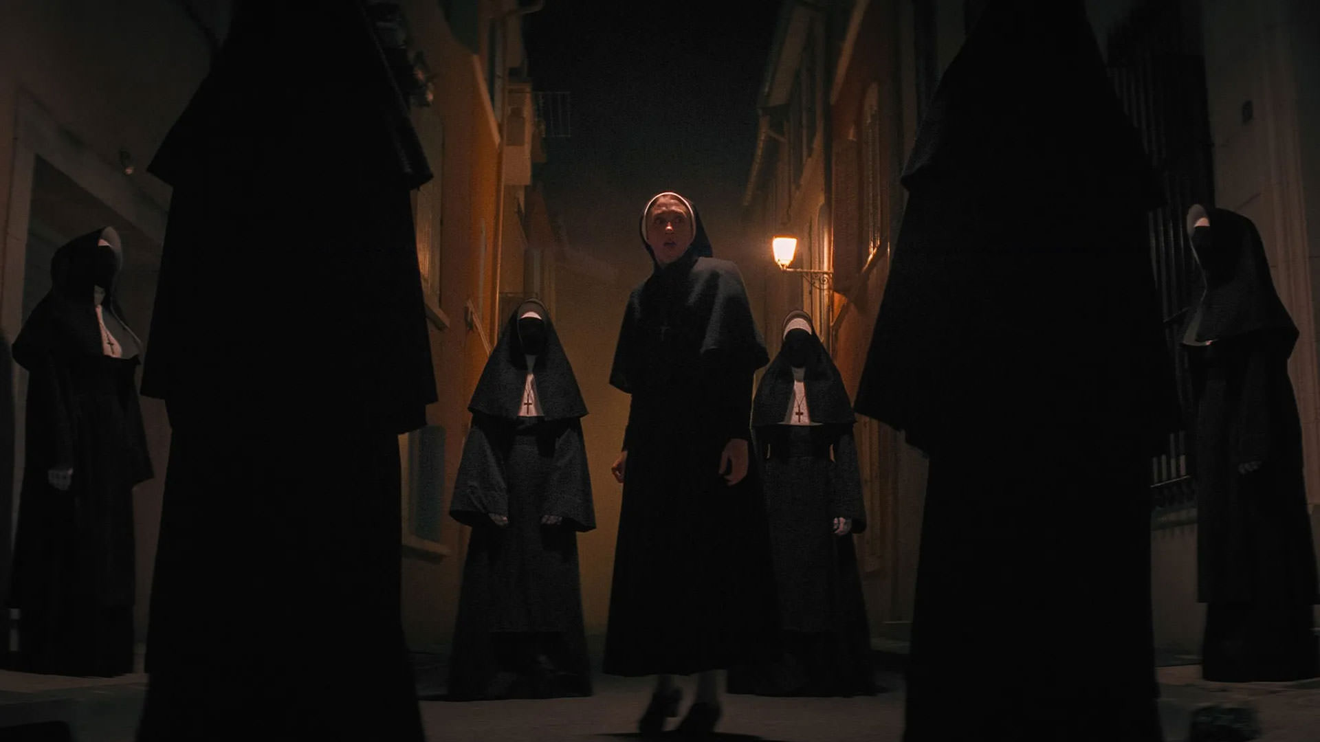 تایسا فارمیگا در کوچه‌ای قدیمی در شب توسط تعدادی راهبه‌ی بی‌چهره احاطه شده است در نمایی از فیلم راهبه ۲ به کارگردانی مایکل چاوز