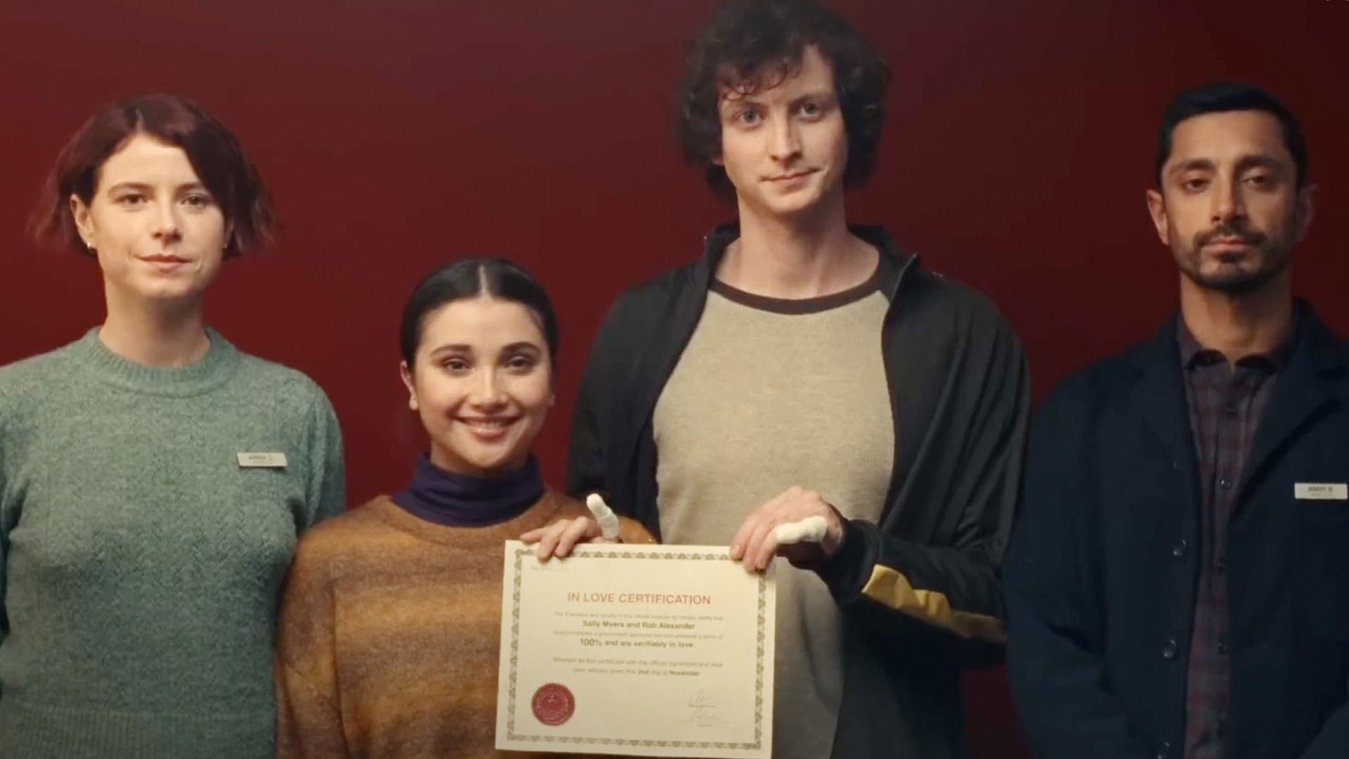 Riz Ahmed et Jesse Buckley, accompagnés de deux autres personnes, prennent une photo de groupe avec un certificat officiel dans une scène du film Nail, réalisé par Christos Niko.