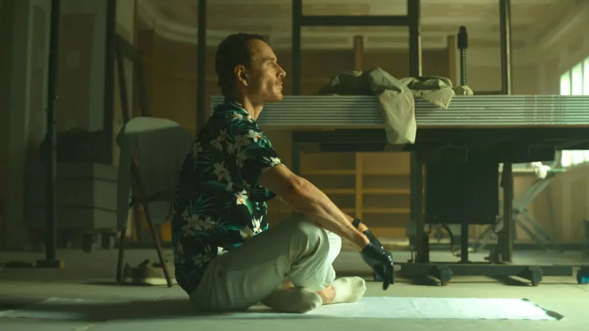 مایکل فاسبندر با پیراهنی طرحدار نشسته وسط یک اتاق نیمه خالی در حالی یوگا کردن در نمایی از فیلم قاتل به کارگردانی دیوید فینچر
