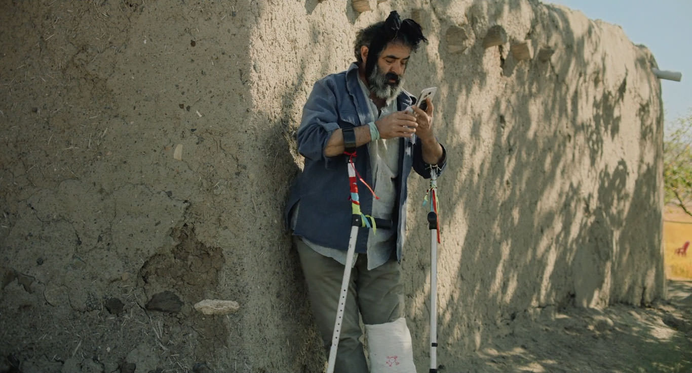 حسن معجونی با پایی شکسته و دو عصا زیر دست ایستاده کنار یک دیوار قدیمی در حال نگاه کردن به گوشی در نمایی از فیلم جاده خاکی به کارگردانی پناه پناهی