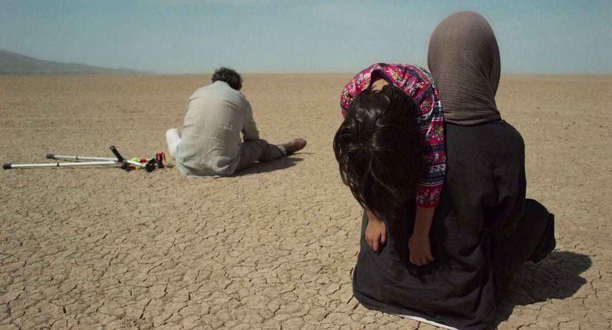 زنی با بچه‌ای بر دوش و مردی با پایی شکسته کمی جلوتر، روی زمین خشک دریاچه‌ی خشکیده‌ی ارومیه نشسته‌اند در نمایی از فیلم جاده خاکی به کارگردانی پناه پناهی