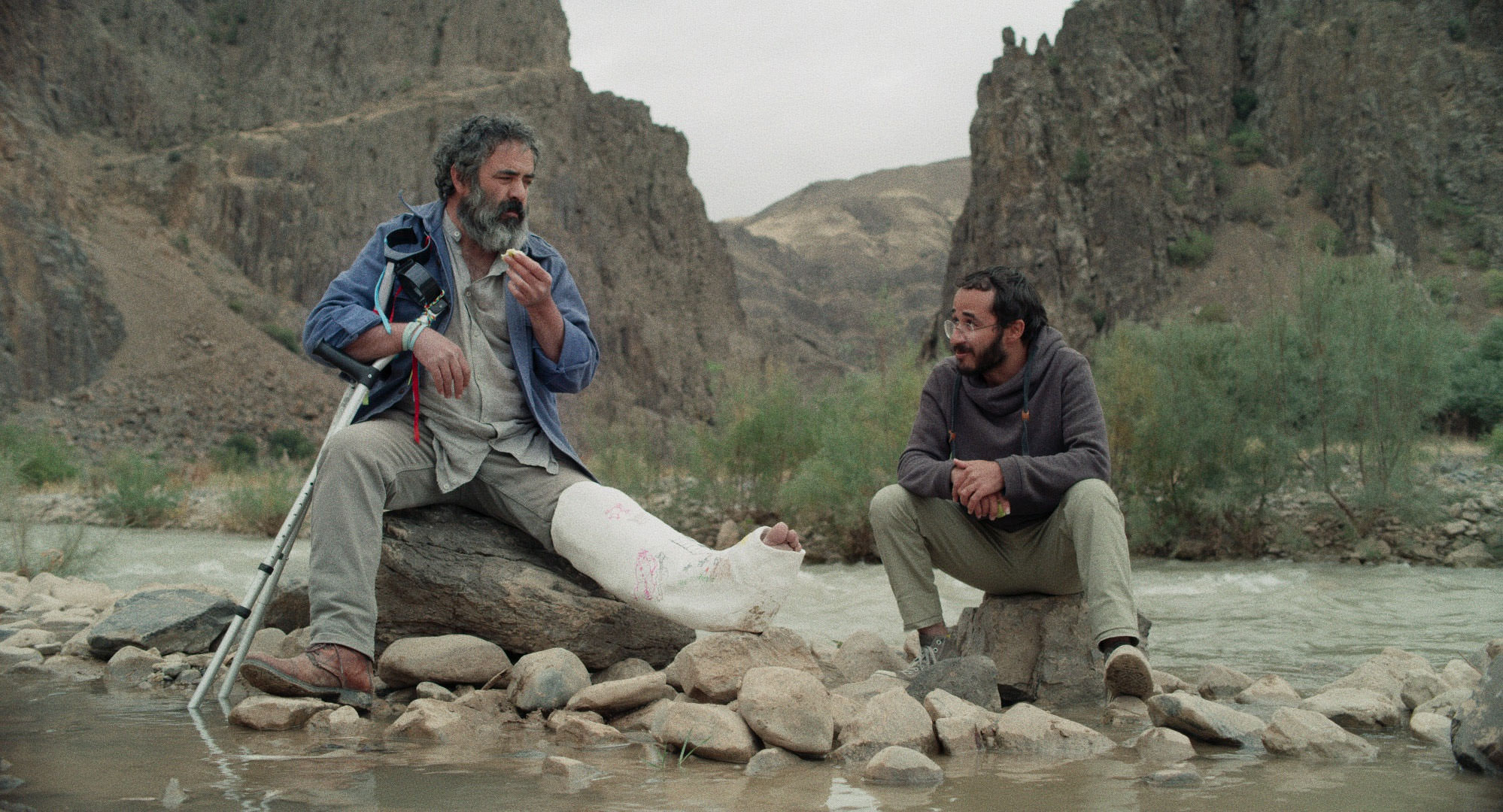امین سیمیار و حسن معجونی با پایی شکسته نشسته کنار یک رود در نمایی از فیلم جاده خاکی به کارگردانی پناه پناهی