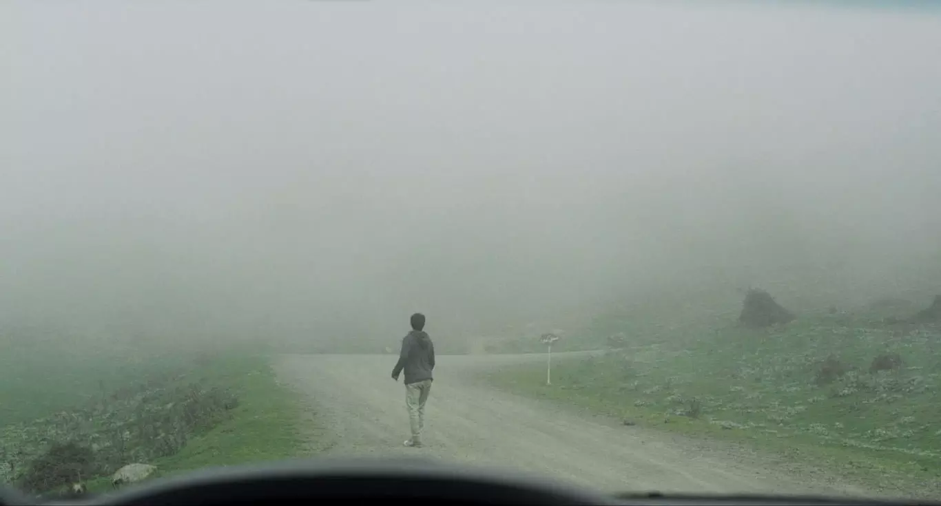 مردی ایستاده در جاده‌ای خاکی در حالی که اطراف‌اش را مه فراگرفته در نمایی از فیلم جاده خاکی به کارگردانی پناه پناهی