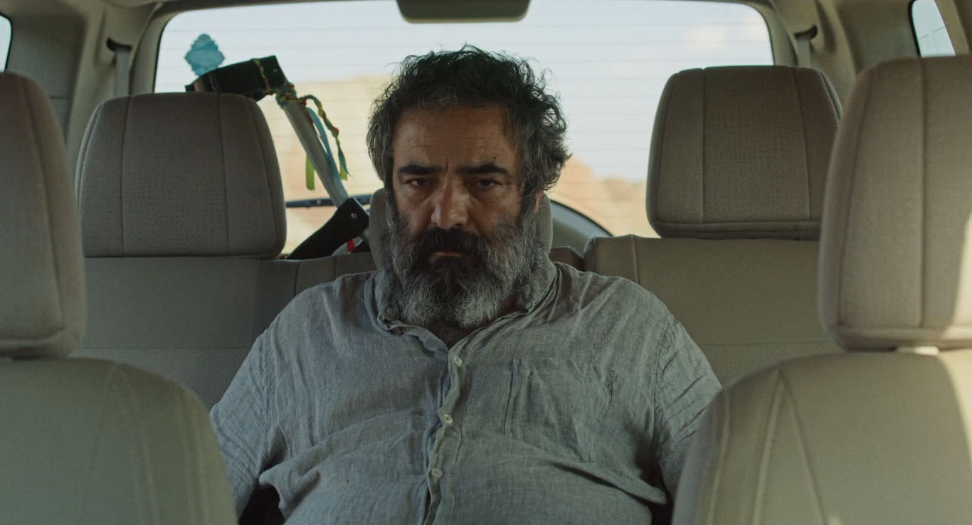 حسن معجونی نشسته روی صندلی عقب یک ماشین در حال نگاه به دوربین در نمایی از فیلم جاده خاکی به کارگردانی پناه پناهی
