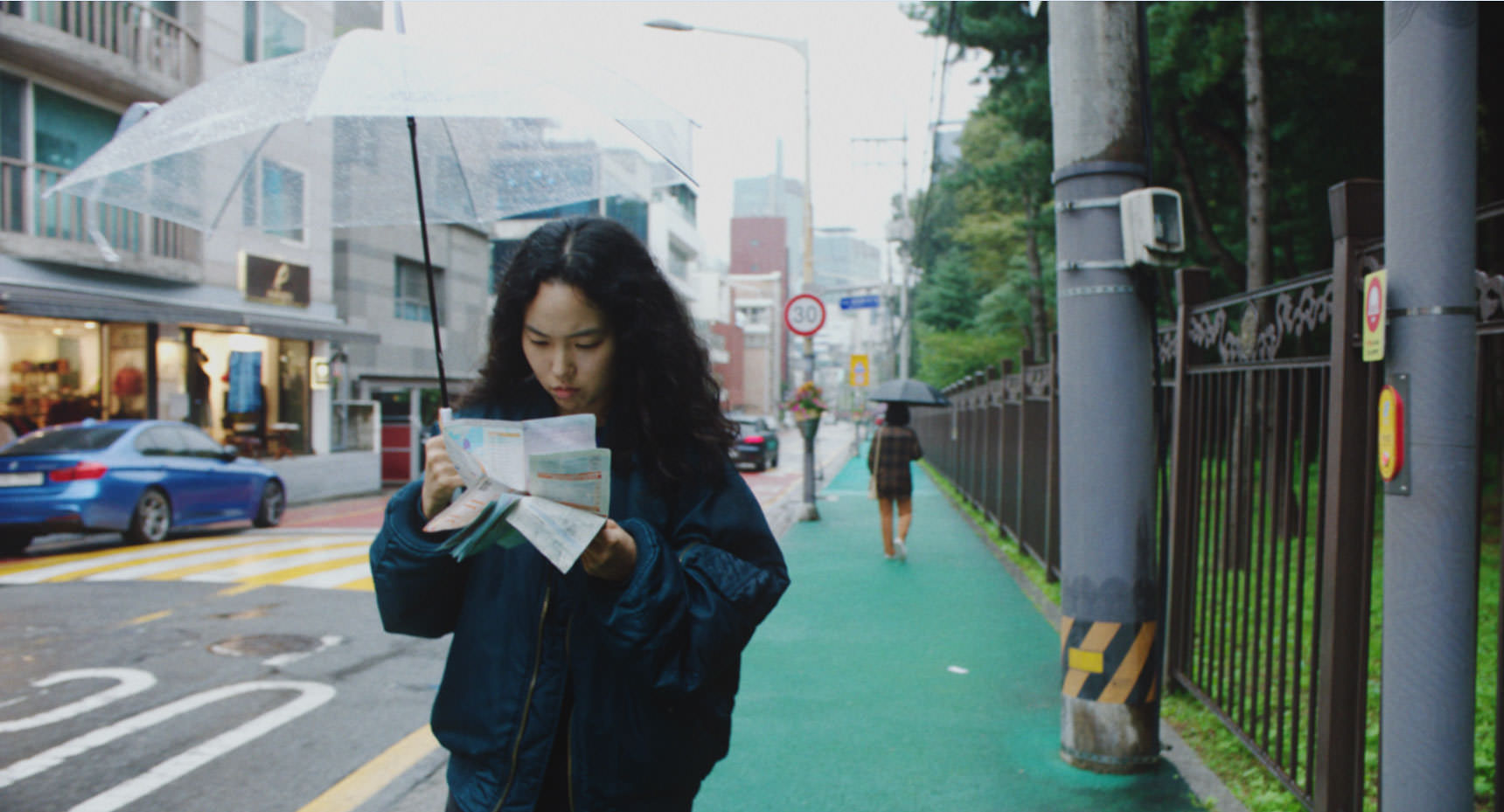 پارک جی-مین در حال مسیریابی با یک نقشه‌ی کاغذی در خیابانی در سئول با چتری در دست در نمایی از فیلم بازگشت به سئول به کارگردانی دیوی چو