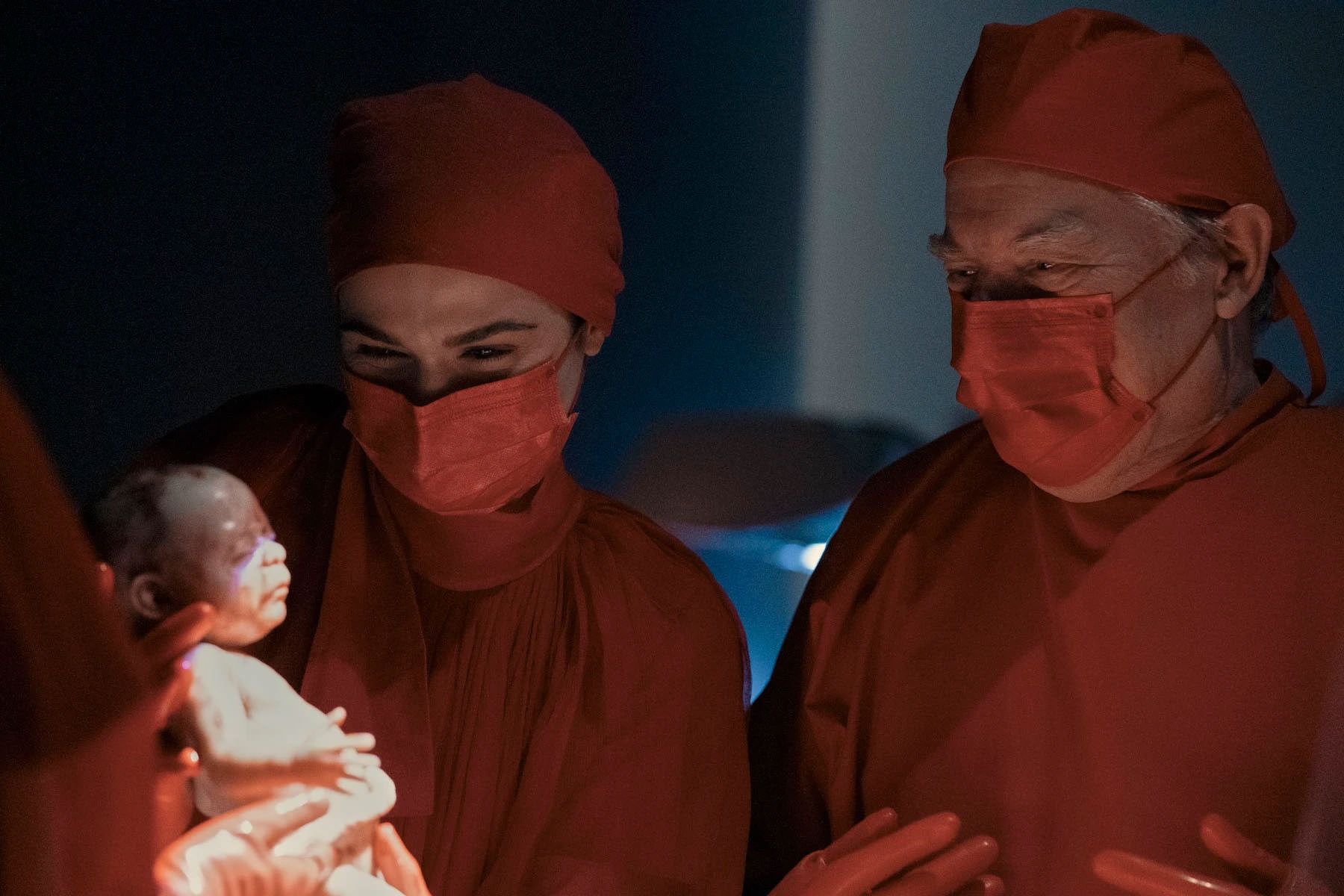 ریچل وایس و مایکل مک‌کین با لباس و ماسک سرخ‌رنگ جراحی در حالی که وایس نوزادی تازه متولد شده را هم در دست دارد در نمایی از سریال شباهت کامل