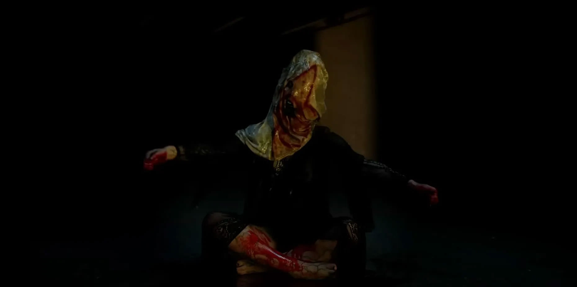 عضوی از یک کالت شیطانی که چهره‌اش با یک کیسه‌ی سفید منقش به طرحی خونین پوشیده شده و روی زمین نشسته و دست‌هایش را باز کرده است در نمایی از فیلم شیطانی به کارگردانی آنتونی دی‌بلاسی