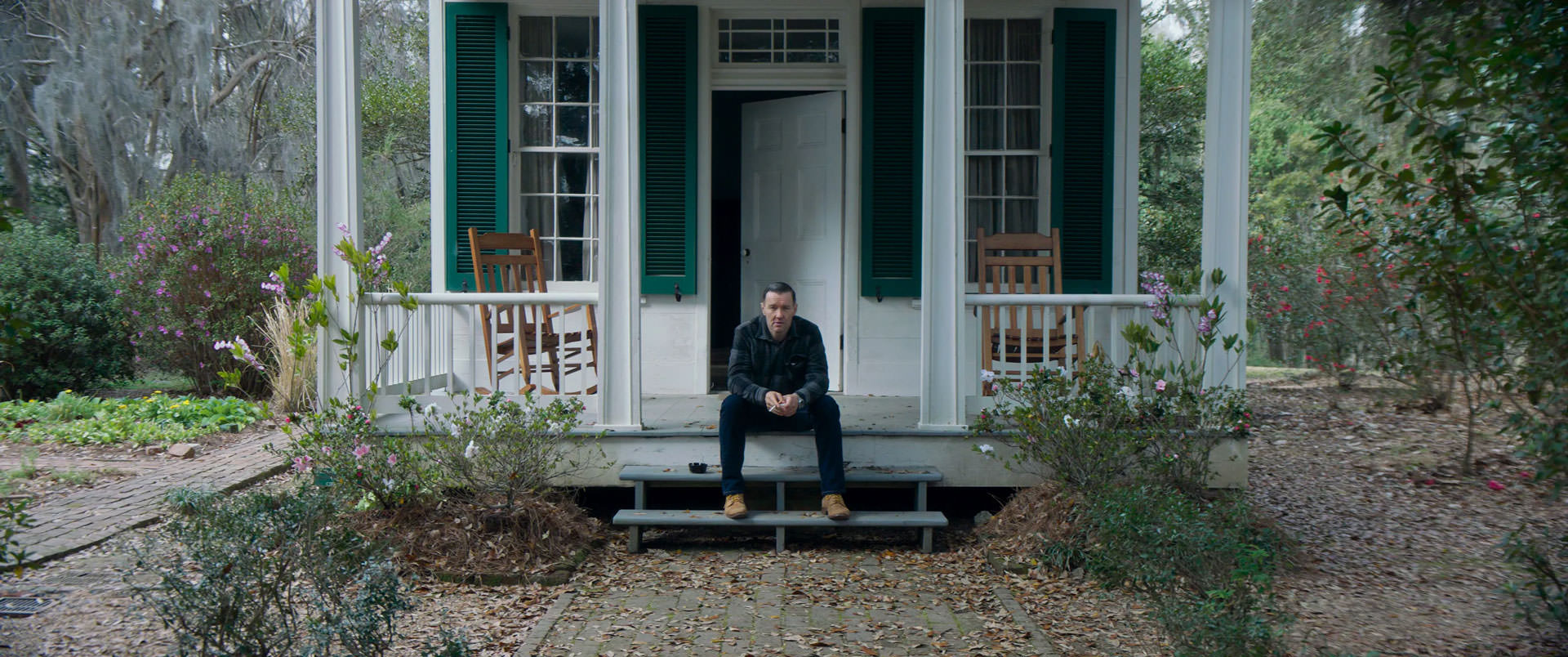 جول اجرتوت نشسته روی پله‌ی مقابل در ورودی یک کلبه‌ی سفید رنگ در نمایی از فیلم استاد باغبان به کارگردانی پل شریدر