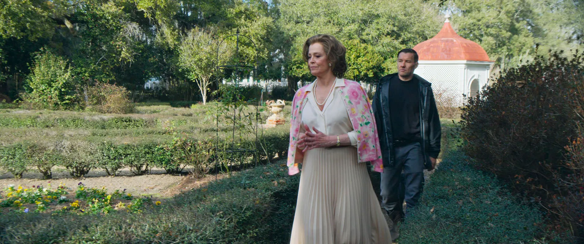 سیگورنی ویور در حال راه رفتن جلوتر در جول اجرتون در یک باغ بازرگ در نمایی از فیلم استاد باغبان به کارگردانی پل شریدر