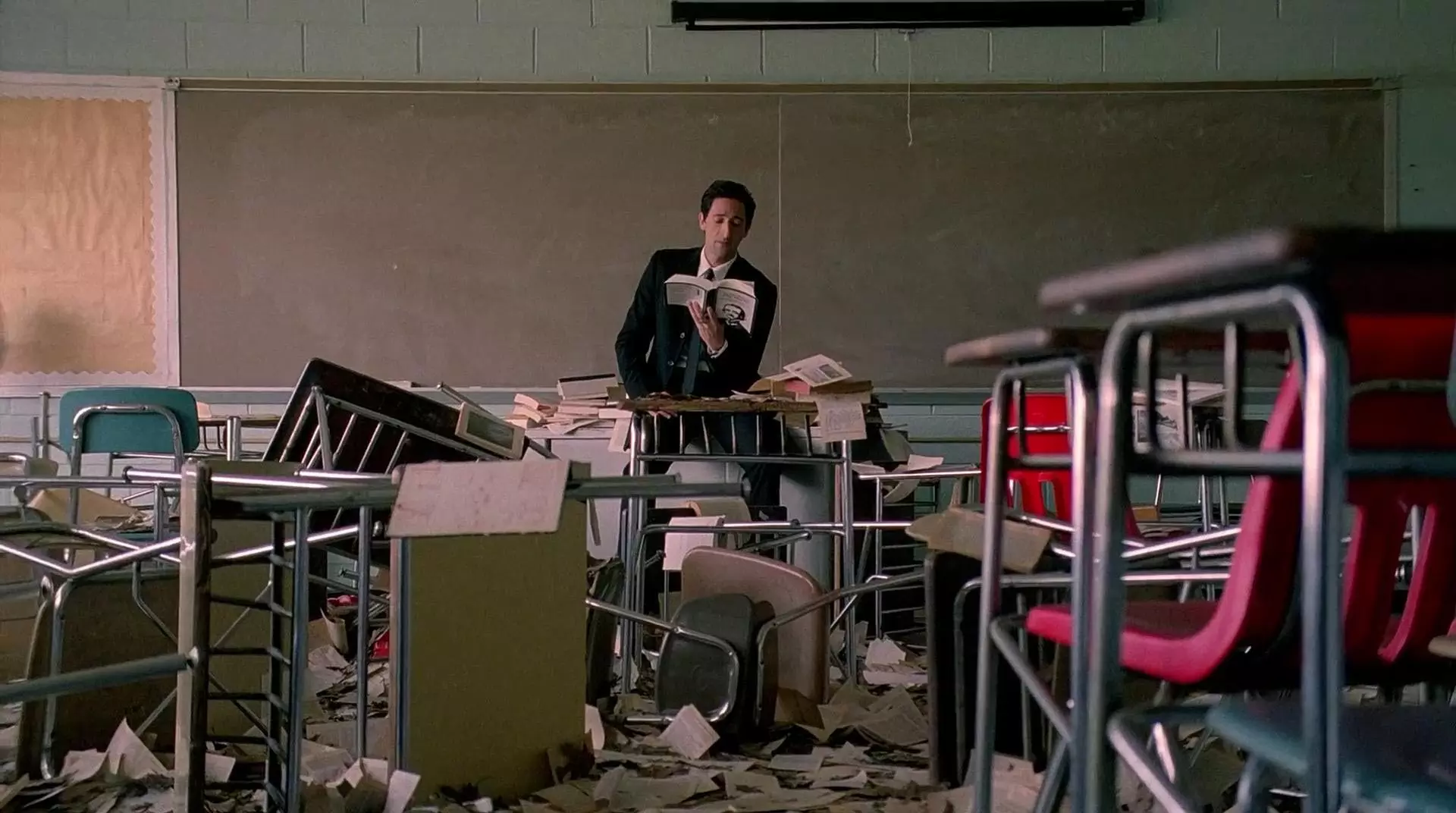 آدرین برودی در میان کلاسی تخریب شده با میز و صندلیهای شکسته در حال خواندن کتاب