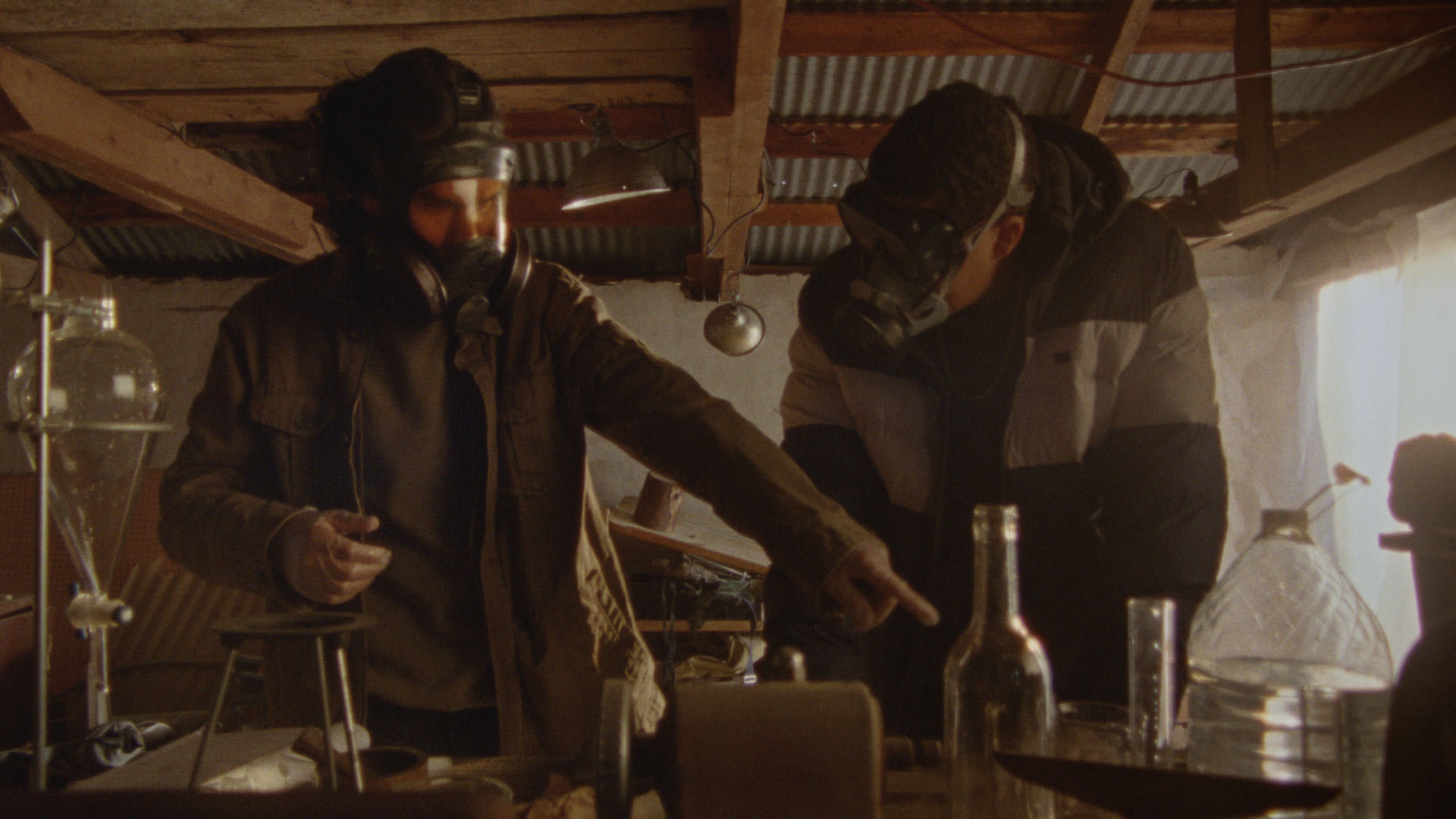 دو مرد با چهره‌هایی که با ماسک محافظ پوشیده شده‌اند در حال ساختن بمب در نمایی از فیلم چگونه یک خط لوله را منفجر کنیم به کارگردانی دنیل گولدهیبر