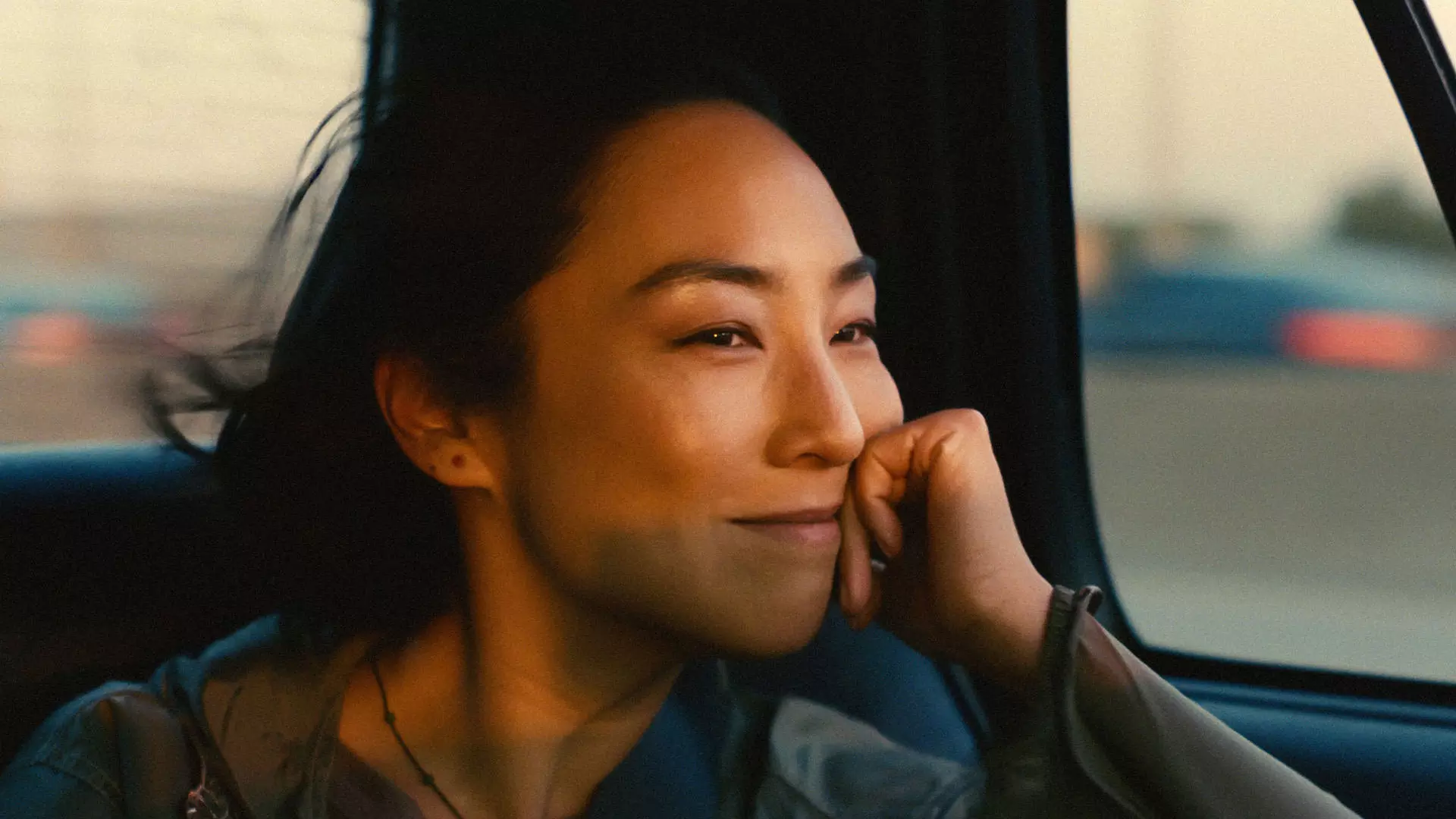 گرتا لی نشسته داخل یک ماشین در حال حرکت در حالی که دستش را زیر چانه‌اش گذاشته و لبخند می‌زند در نمایی از فیلم زندگی‌های پیشین به کارگردانی سلین سانگ