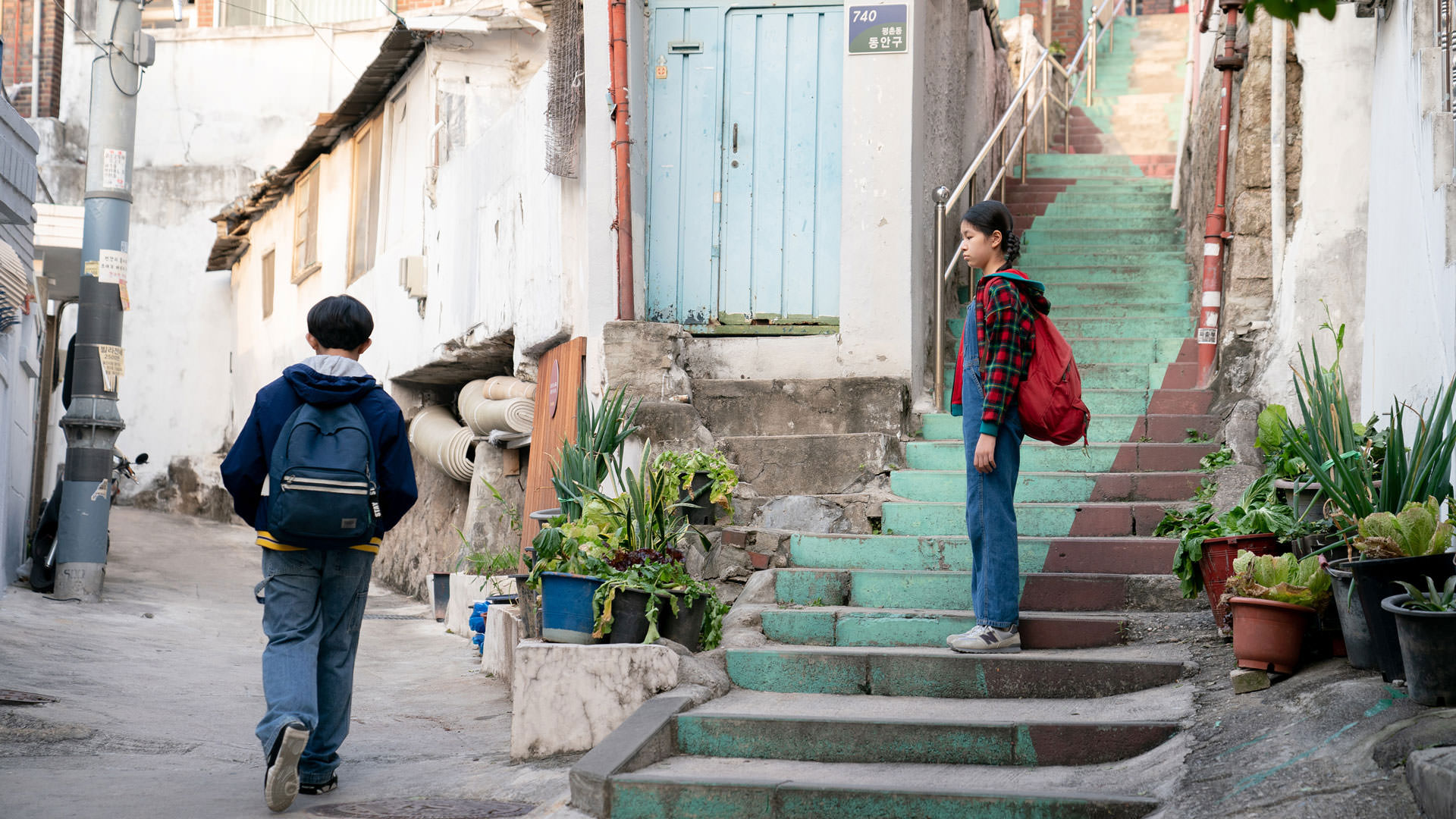دختربچه‌ای که روی پله‌ها ایستاده است قدم زدن یک پسربچه در کوچه را تماشا می‌کند در نمایی از فیلم زندگی‌های پیشین به کارگردانی سلین سانگ
