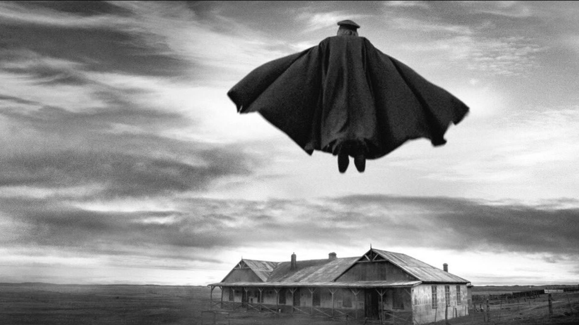 مردی با لباس نظامی و یک شنل بلند بر فراز خانه‌ای واقع در یک دشت پرواز می‌کند در نمایی سیاه و سفید از فیلم کنت به کارگردانی پابلو لارائین