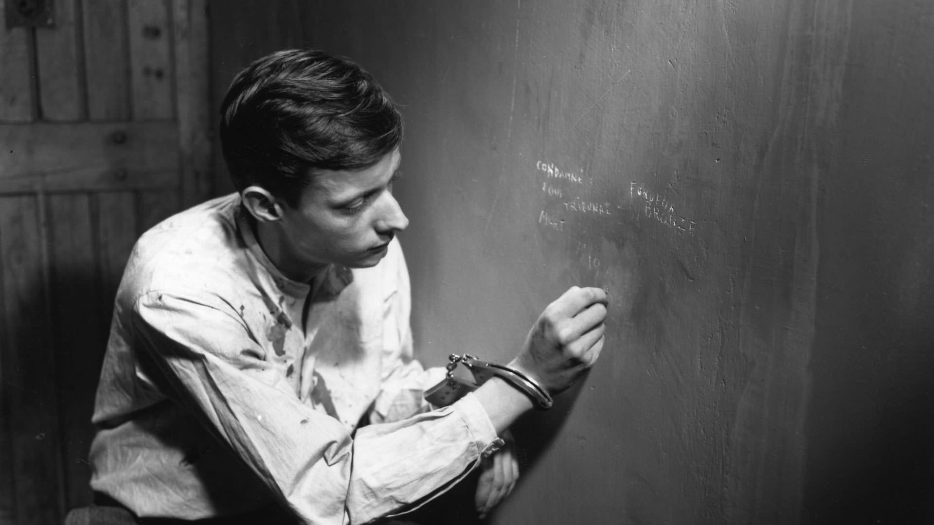 فرانسوا لتریه با دستانی دستبند زده در حال نوشتن روی دیوار یک سلول انفرادی در نمایی از فیلم مردی گریخت به کارگردانی روبر برسون