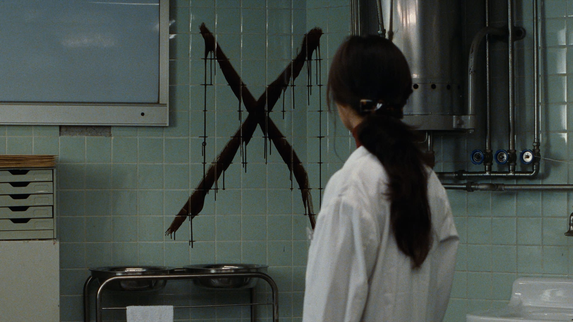 زنی با کت سفید دکتر در صحنه ای از فیلم درمان کیوشی کوروساوا به صلیب سیاه روی دیوار نگاه می کند.