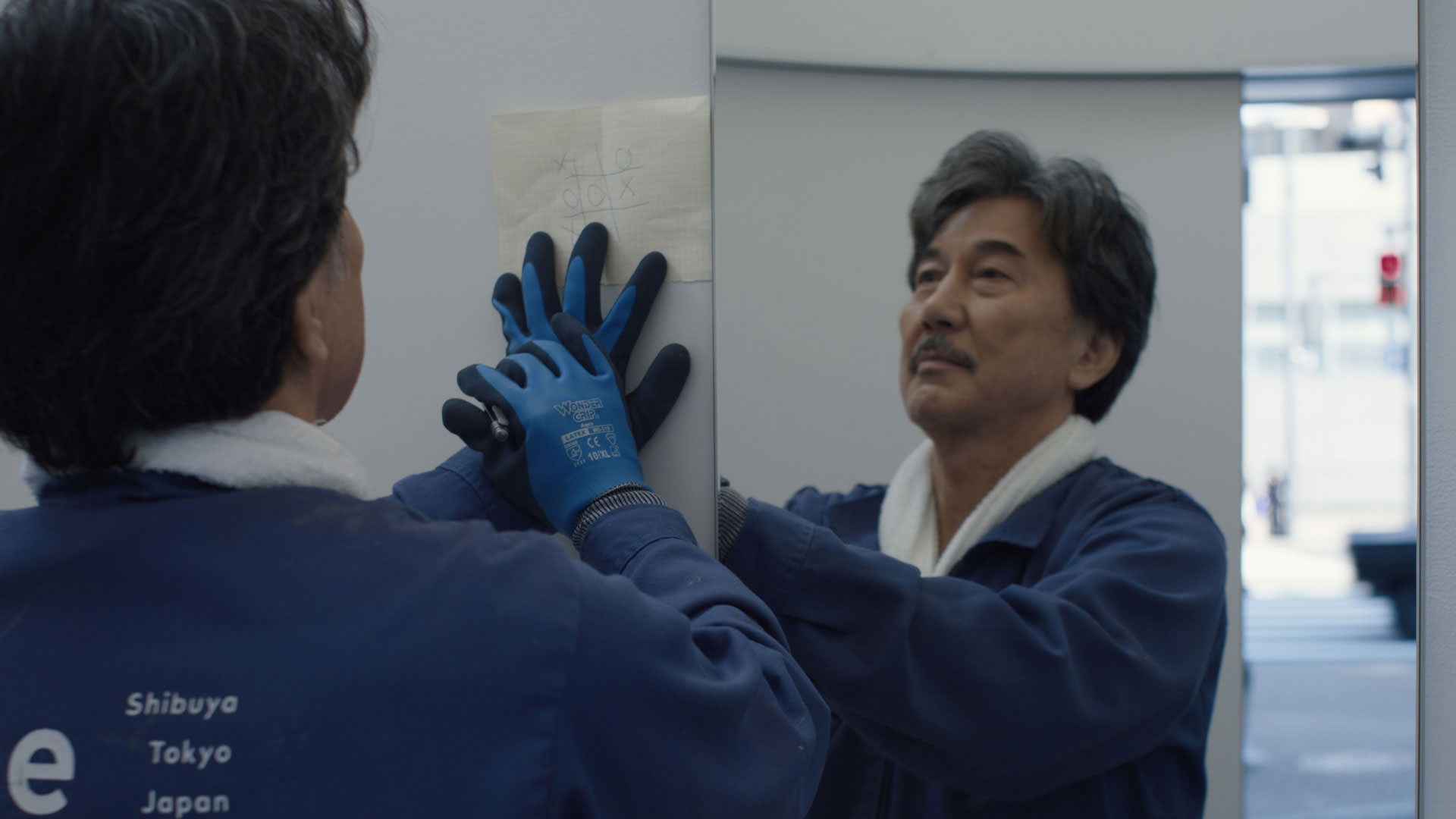 کوجی یاکوشو با لباس فرم آبی در حال تمیز کردن آینه‌ی یک سرویس بهداشتی عمومی در نمایی از فیلم روزهای عالی به کارگردانی ویم وندرس