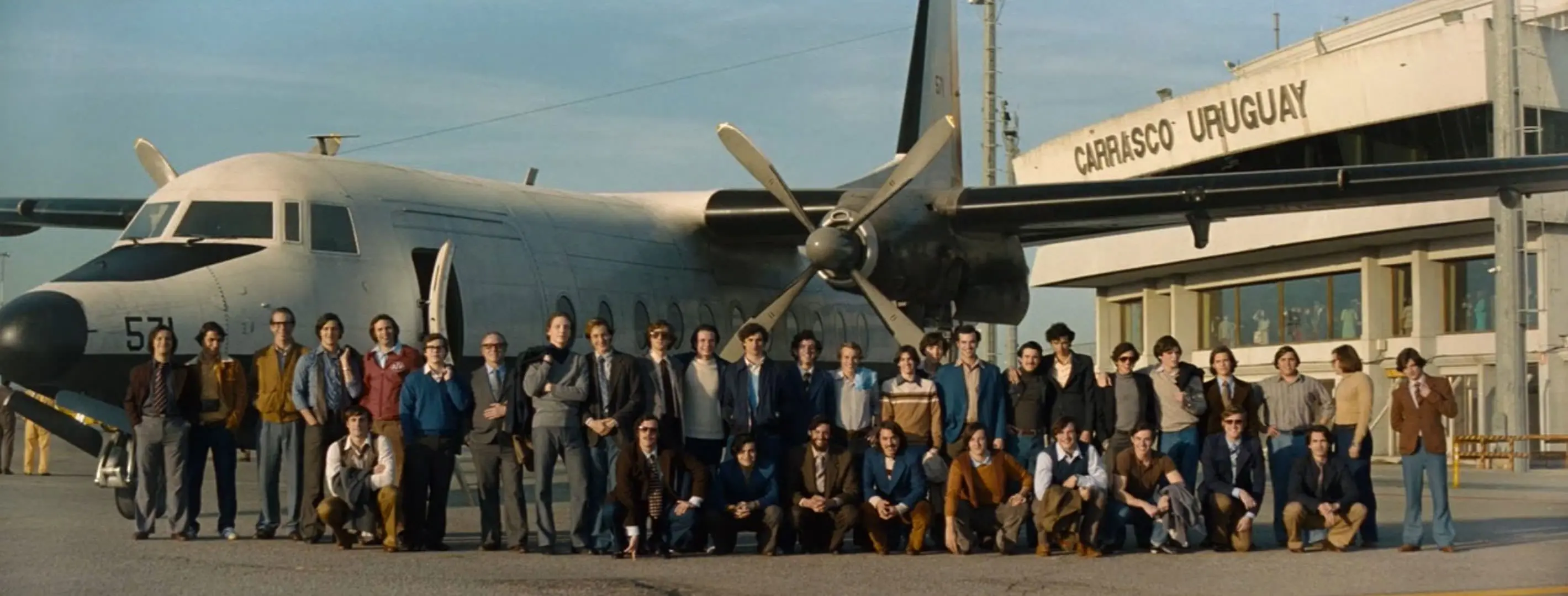   Vol 571 de l'armée de l'air uruguayenne avec des passagers debout devant l'avion à l'aéroport et prenant une photo de groupe commémorative dans une scène du film Snow Society réalisé par Jay Bayona. 