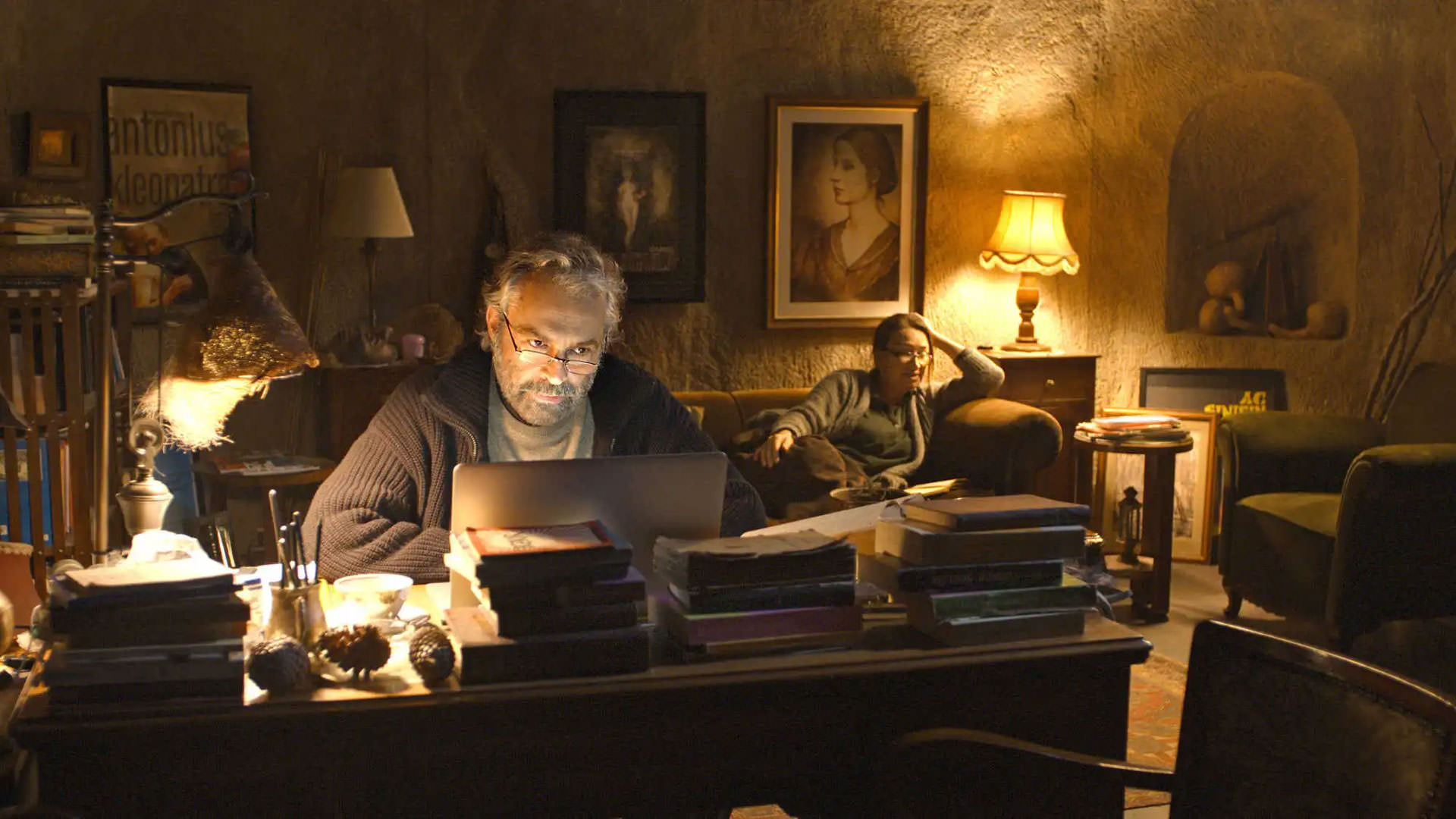 هالوک بیلگینر پشت میز می نشیند و با لپ تاپ در اتاقی با نور زرد رنگ می نویسد در حالی که دمت اکبا پشت سر او روی کاناپه نشسته است در صحنه ای از فیلم خواب زمستانی به کارگردانی نوری بیلگه جیلان.