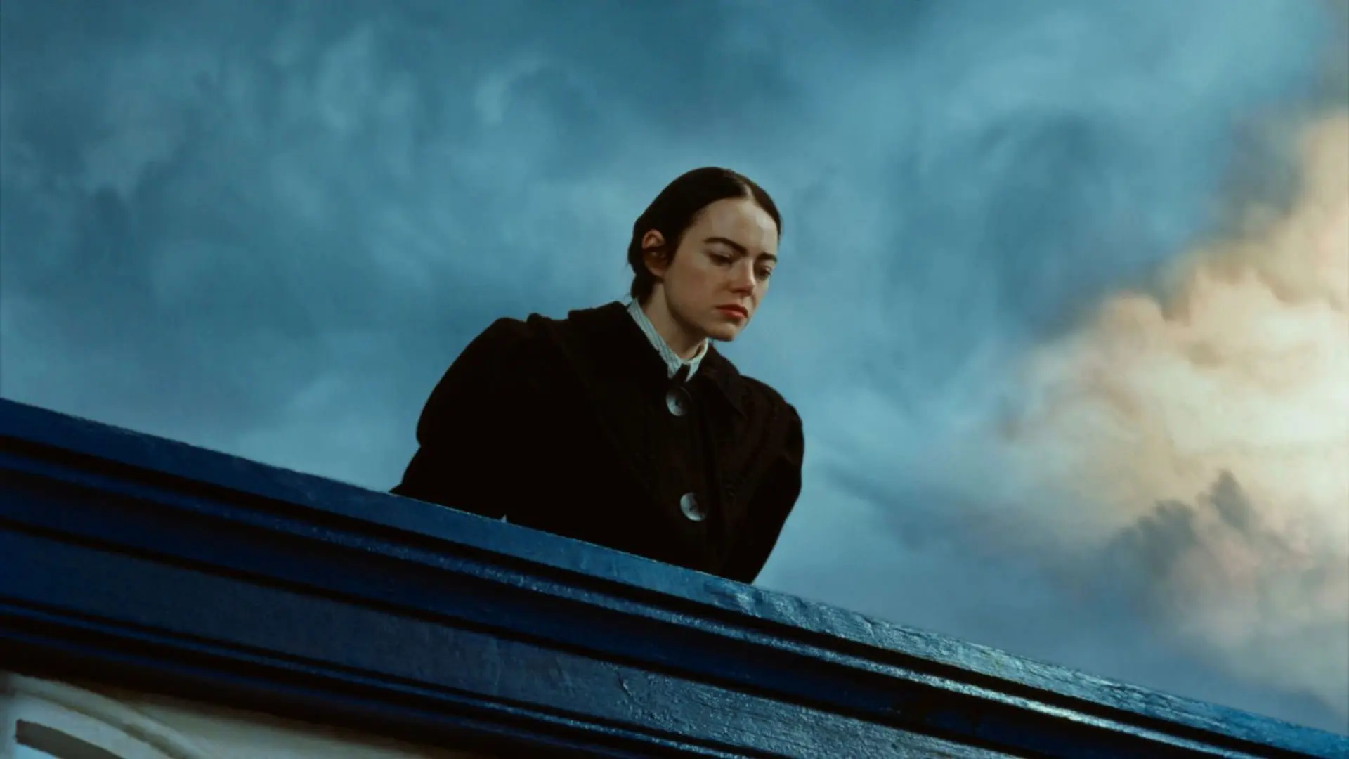 اما استون با لباس مشکی از بالای یک پل به پایین آن نگاه می‌کند در حالی که آسمان آبی و ابری پشت سرش دیده می‌شود در نمایی از فیلم بیچارگان به کارگردانی یورگوس لانتیموس