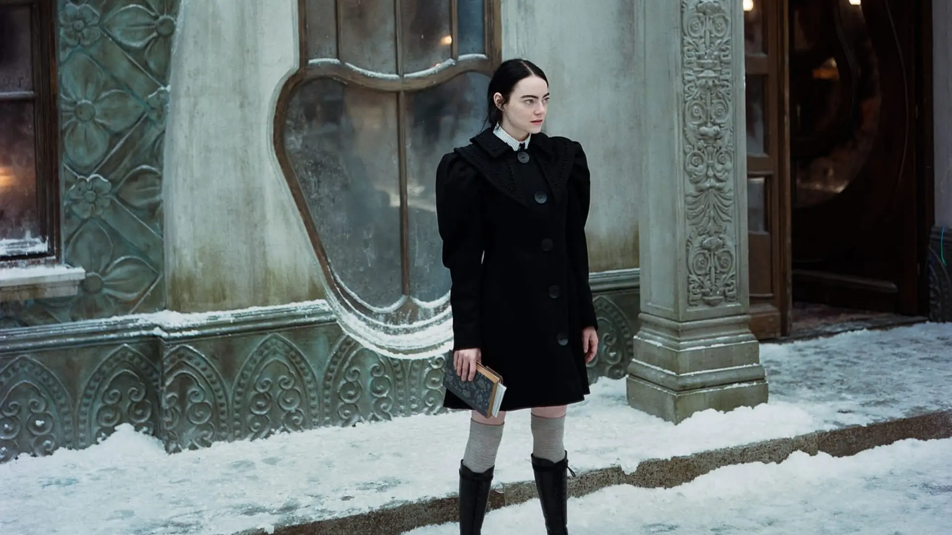 اما استون با لباس مشکی ایستاده مقابل یک ساختمان در حالی که زمین برفی است در نمایی از فیلم بیچارگان به کارگردانی یورگوس لانتیموس