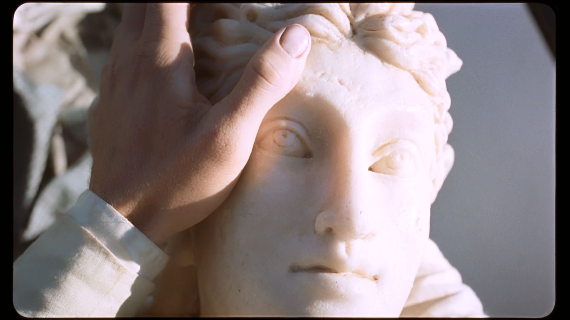 دستان مردی سفیدپوش روی صورت یک مجسمه‌ی سفید قرار گرفته است که آفتاب روی آن تابیده در نمایی از فیلم کیمرا به کارگردانی آلیچه رورواکر