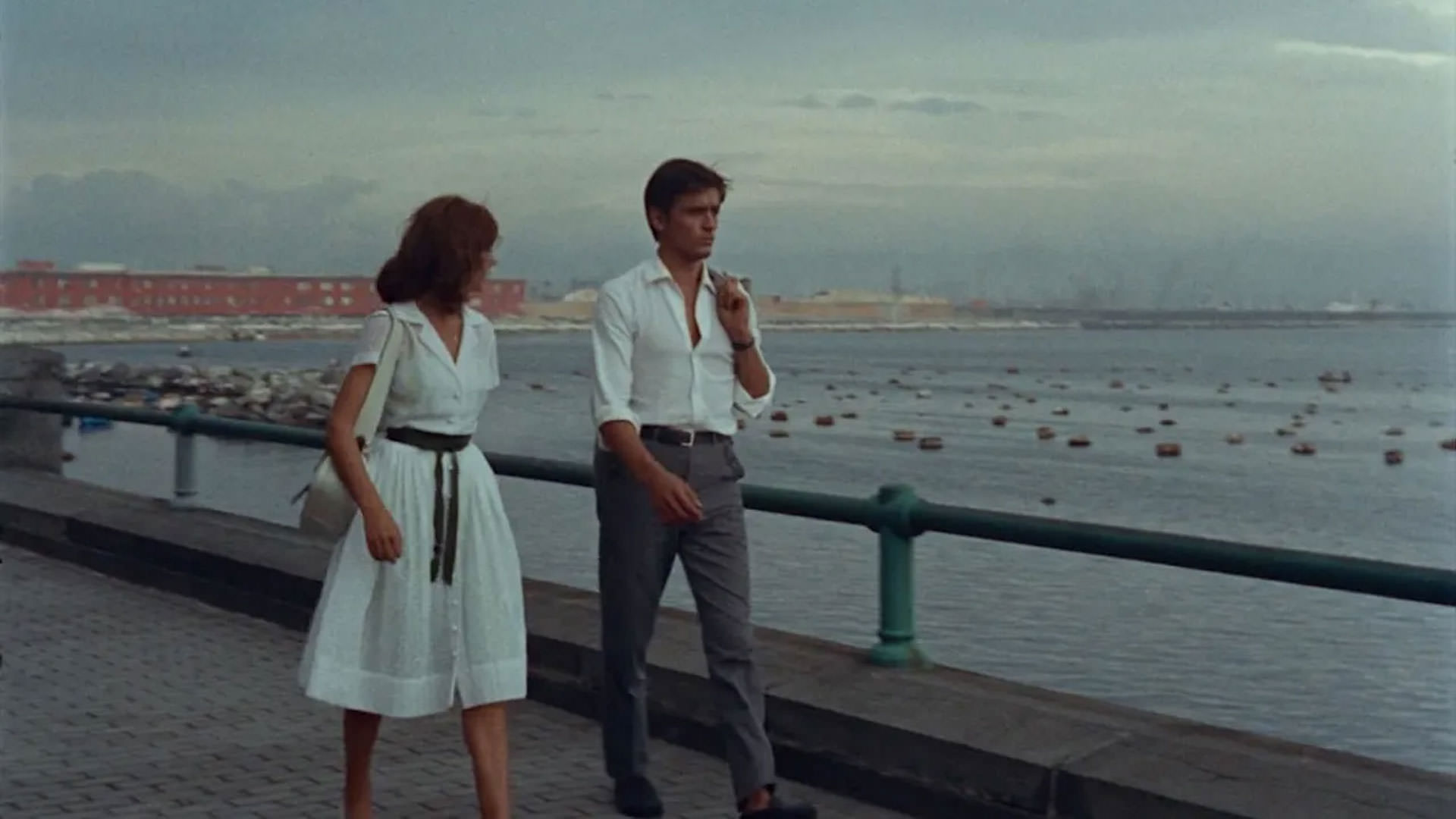 آلن دلون و ماری لافوره با لباس‌هایی سفید در حال قدم زدن کنار دریا در نمایی از فیلم ظهر بنفش به کارگردانی رنه کلمان