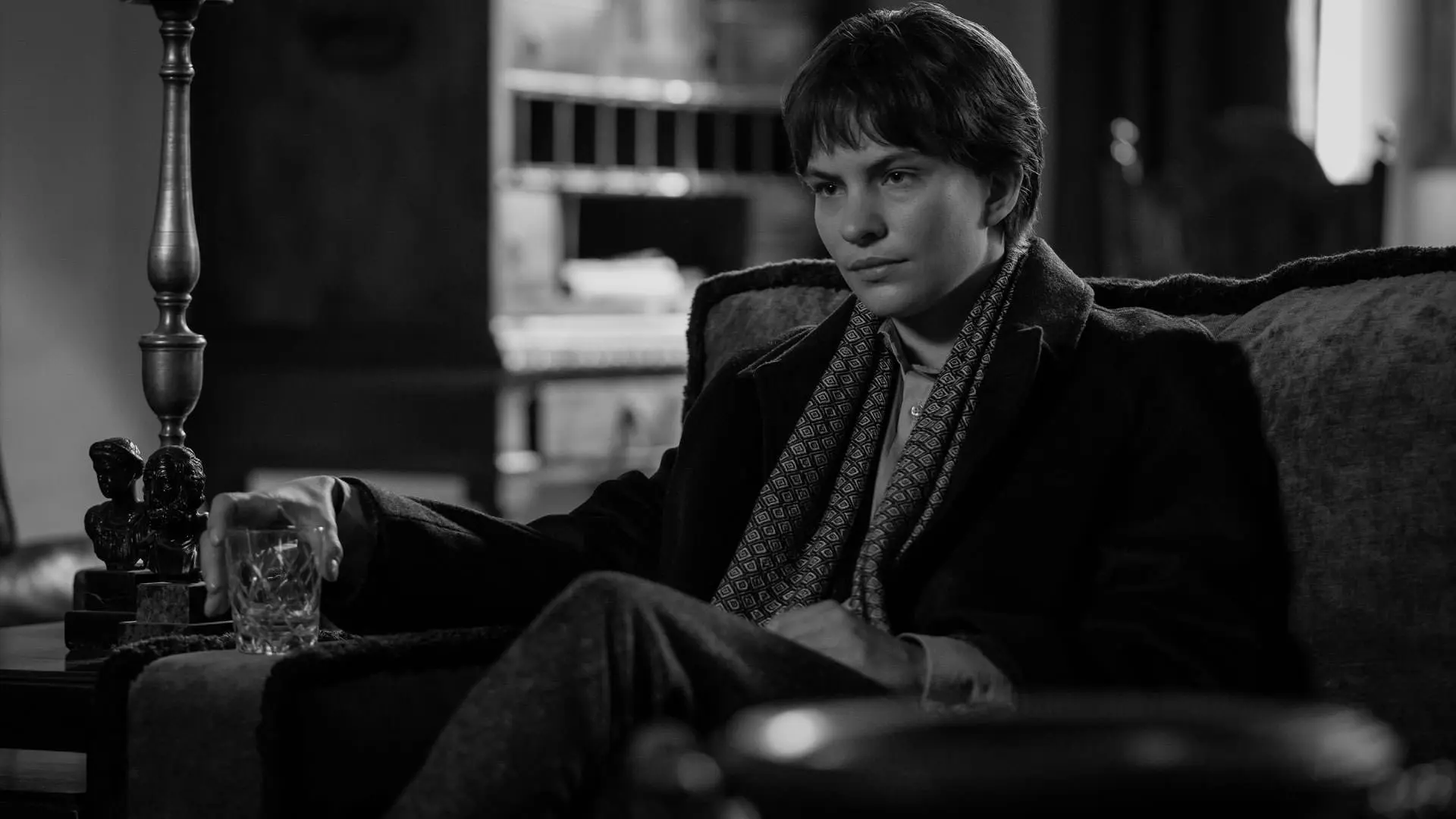 الیوت سامر با یک لیوان در دست در یک عکس سیاه و سفید از سریال ریپلی استیون زیلیان نشسته است.