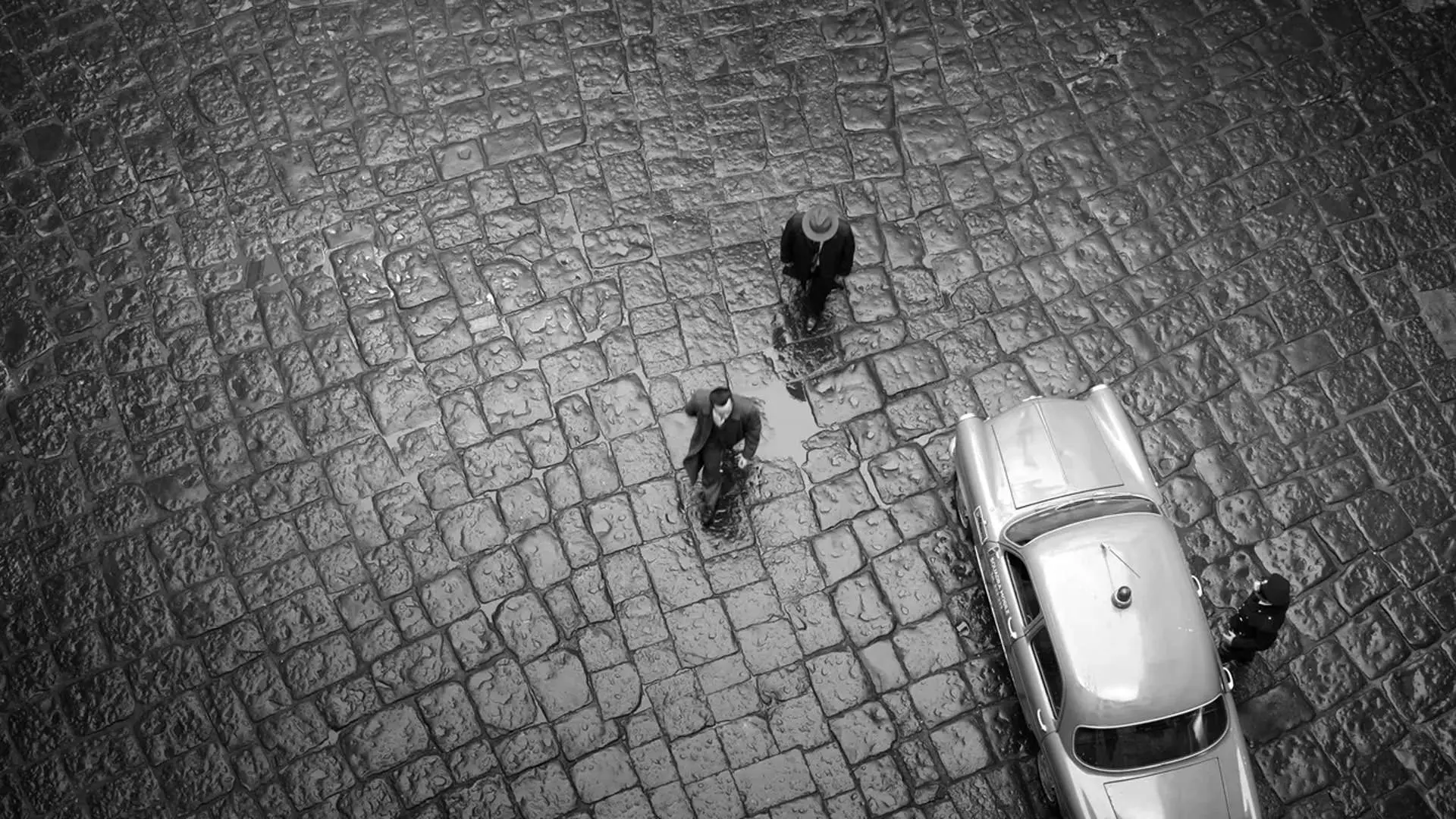 نمای سیاه و سفید بالای سر دو مرد در حال راه رفتن در کنار ماشین در سریال ریپلی به کارگردانی استیون زیلیان.