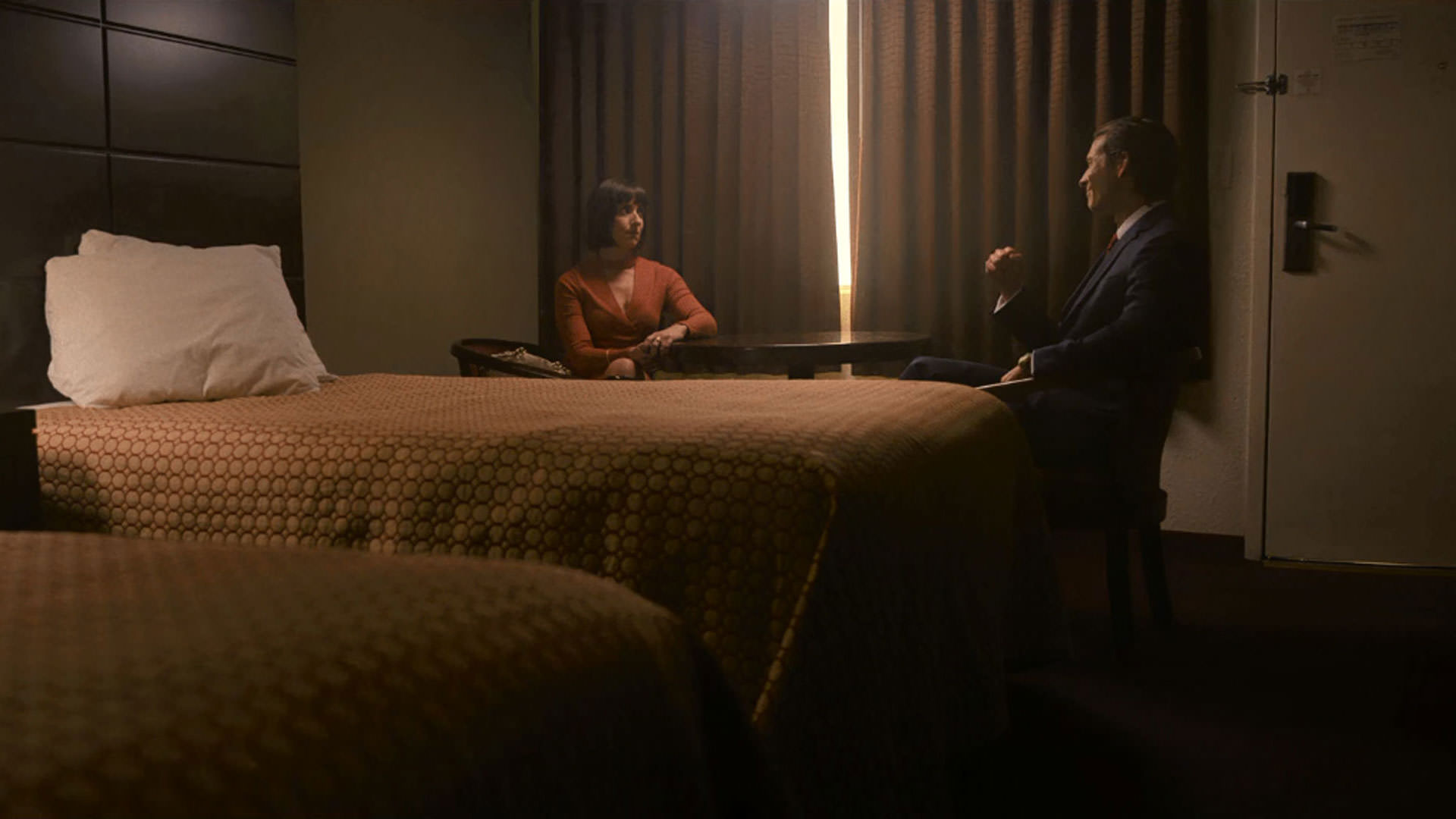 گلن پاول نشسته مقابل یک زن داخل اتاق نیمه‌خالی و تاریک یک متل در نمایی از فیلم آدمکش به کارگردانی ریچارد لینکلیتر
